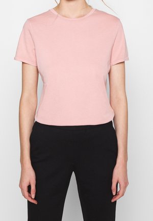 Persona que lleva una camiseta lisa de manga corta color rosa claro y pantalones negros, mostrada desde el cuello hasta las caderas sobre un fondo blanco.