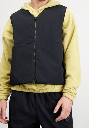 Osoba nosící černou vestu na zip přes žlutou fleecovou mikinu s kapucí a černé kalhoty Nike, stojící s rukama uvolněně podél těla.