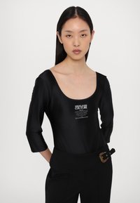 Body negro de manga larga en tejido suave con escote amplio y detalle de logo impreso. Combinado con pantalones negros de cintura alta que presentan un cinturón con acento dorado.