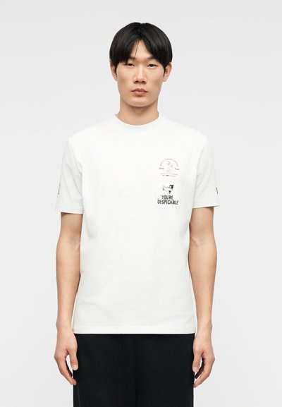 Jeune homme aux cheveux noirs courts portant un t-shirt blanc avec de petits patchs graphiques et du texte, debout devant un fond clair uni.