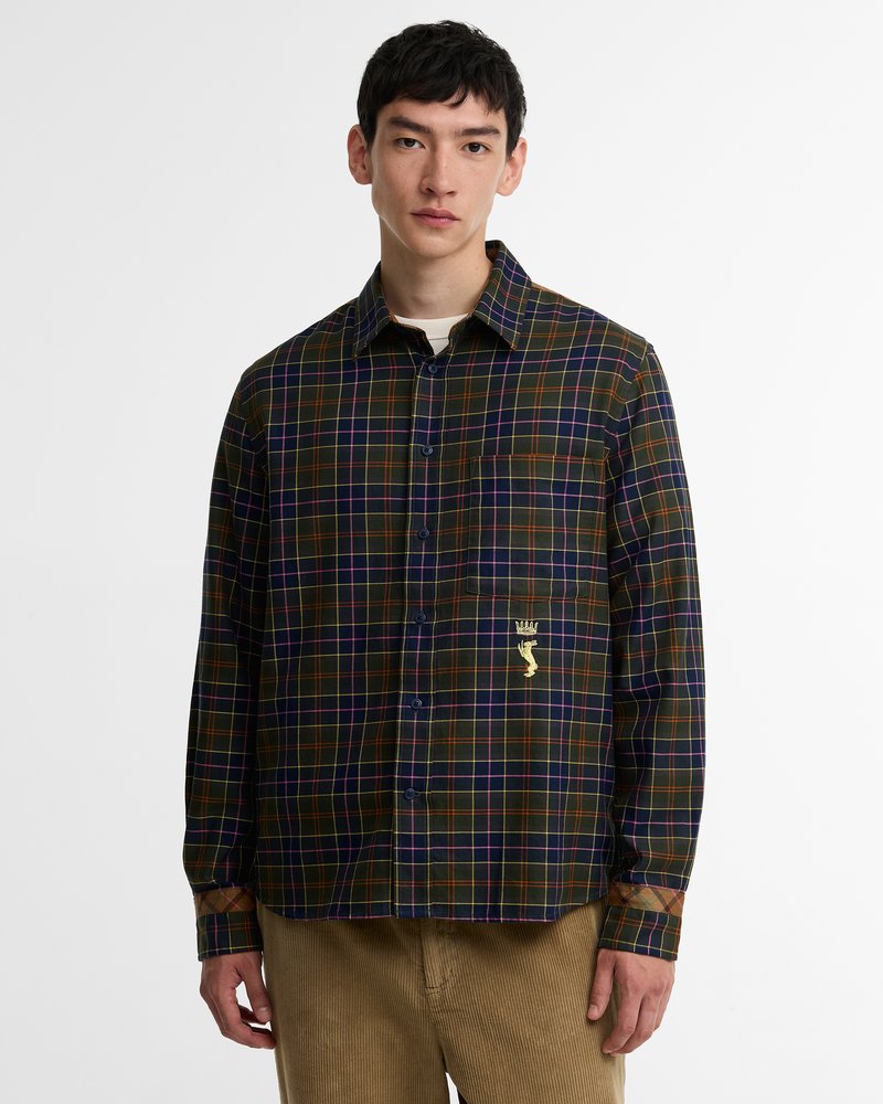 Jeune homme portant une chemise à carreaux foncés boutonnée, avec une poche frontale et un détail brodé d'une couronne dorée et d'une main, associée à un pantalon marron clair.