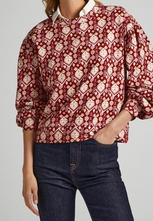 Kobieta ubrana w czerwoną i beżową bluzkę z długim rękawem w wzory, nałożoną na białą koszulę z kołnierzykiem, w ciemnoniebieskich dżinsach, z ręką w kieszeni.