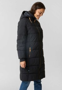 Woman smiling while wearing a long black quilted winter coat with a hood, zipped pockets, and blue jeans against a plain background.