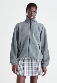 Graue Fleecejacke mit hohem Kragen, Reißverschluss und Seitentaschen. Getragen über einem schwarz-weißen karierte Faltenrock.