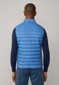 Blå puffväst med horisontell quiltning, hög krage och ärmlös design, bärs över en mörk tröja. Slät textur och lättviktig material.