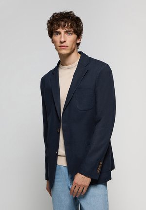 Blazer bleu marine avec poche sur la poitrine, deux boutons et tissu texturé, associé à une chemise beige et un jean bleu clair.