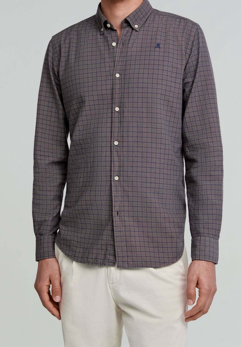 Homme portant une chemise grise à carreaux à manches longues, boutonnée avec col, et un pantalon blanc sur un fond clair uni.