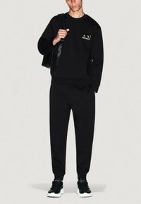 Joven con sudadera negra con el logo de AX, joggers negros a juego, zapatillas en blanco y negro, y llevando un bolso negro cruzado.