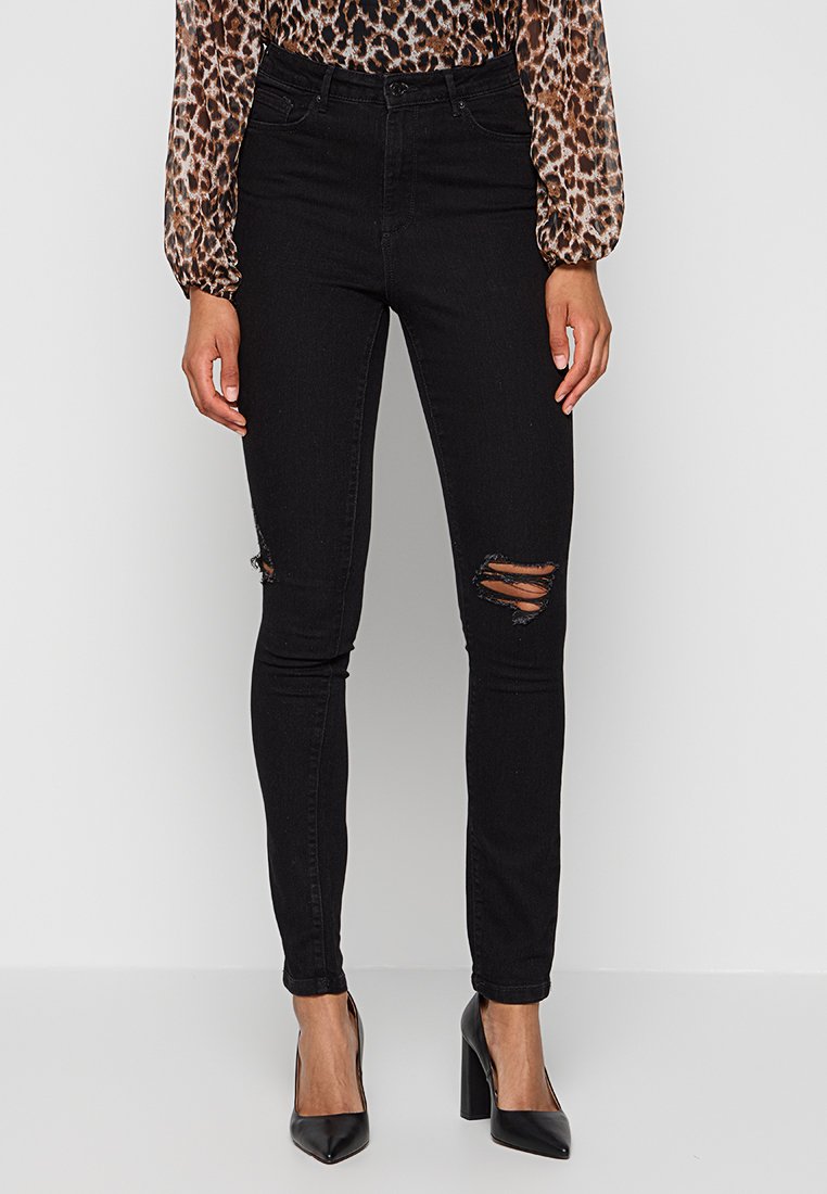 Svarta smala jeans med slitna hål vid knäna, tillverkade av denimmaterial. Designade med en åtsittande passform och parat med svarta högklackade skor.