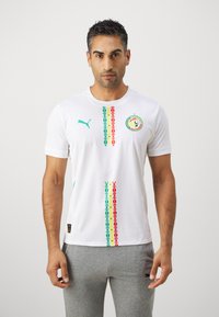 Bela majica s kratkimi rokavi z barvitimi navpičnimi vzorci v rdeči, zeleni in rumeni barvi. Ima okrogel izrez in Puma logotip v teal barvi.