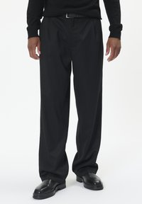 Čierne plisované nohavice z hladkej tkaniny, s voľným strihom, doplnené lesklými čiernymi topánkami.