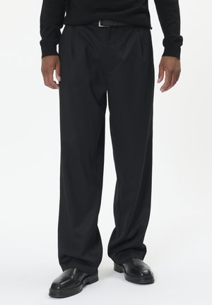 Pantalons plissés noirs en tissu à texture lisse, dotés d'une coupe décontractée, associés à des chaussures noires brillantes.