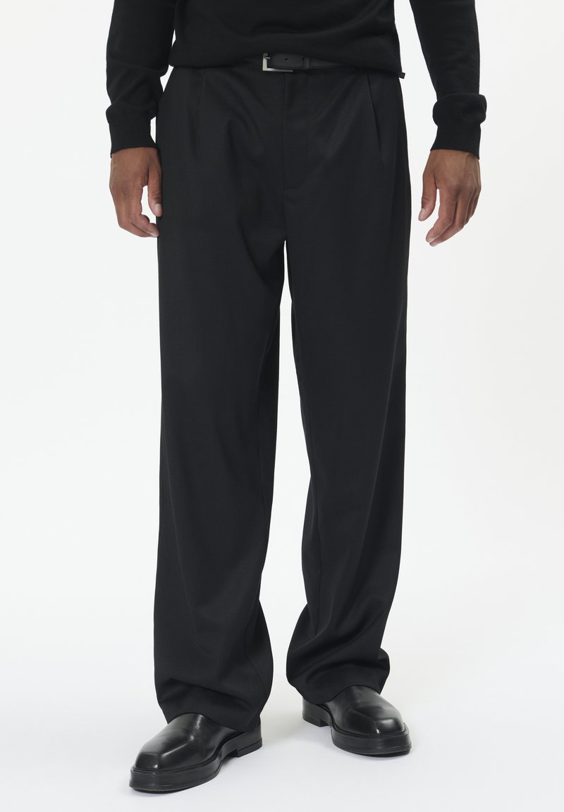 Čierne plisované nohavice z hladkej tkaniny, s voľným strihom, doplnené lesklými čiernymi topánkami.