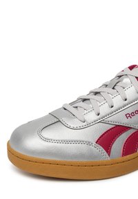 Srebrne skórzane sneakersy z czerwonymi akcentami, gumową podeszwą, okrągłym noskiem i płaskimi sznurowadłami. Posiadają widoczne przeszycia i teksturowaną powierzchnię.