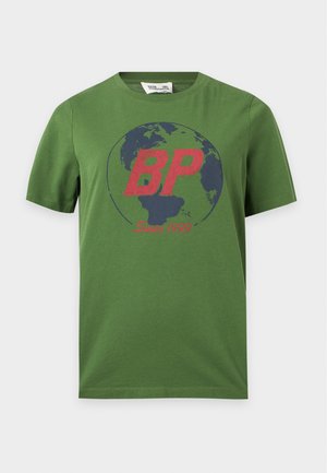 Femme aux longs cheveux foncés portant un t-shirt vert avec un motif graphique et un jean large bleu clair, debout avec une main dans la poche.