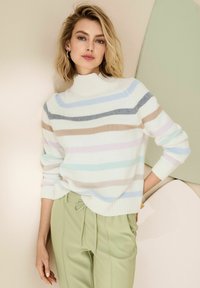 Vrouw met schouderlang blond haar, gekleed in een witte coltrui met pastelstrepen en lichtgroene broek met koordjes, poseert binnenshuis.