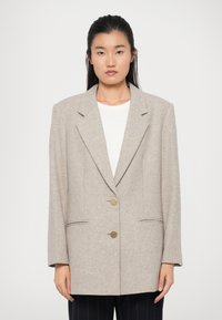 Beige herringbone blazer with notched lapels, two gold-tone buttons, and side pockets. Oversized fit with textured fabric.