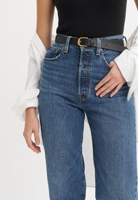 Personne portant un jean bleu taille haute avec une ceinture noire à boucle dorée, un haut noir sans manches et une chemise blanche à manches longues drapée sur les bras.