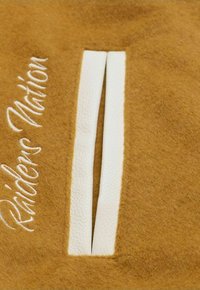 Brown felt fabric with white leather accents featuring two parallel cutouts. Embroidered text "Raiders Nation" in white.