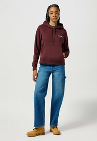 Bordeauxkleurige hoodie met kangoeroezak en trekkoord; gecombineerd met wijdachtige blauwe jeans en tan kleurige werklaarzen.