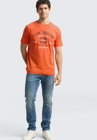 Pomarańczowy bawełniany T-shirt z krótkimi rękawami, z nadrukiem "TOM TAILOR" w ciemnoniebieskim kolorze, zestawiony z jasnoniebieskimi jeansami i białymi trampkami.