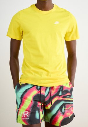 Homme portant un T-shirt Nike jaune vif et un short Nike multicolore à motif abstrait, les mains dans les poches, sur un fond uni.
