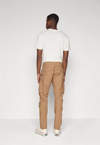 Man die met zijn rug naar de camera staat, gekleed in een witte T-shirt met korte mouwen, beige cargobroek met meerdere zakken en witte high-top sneakers.