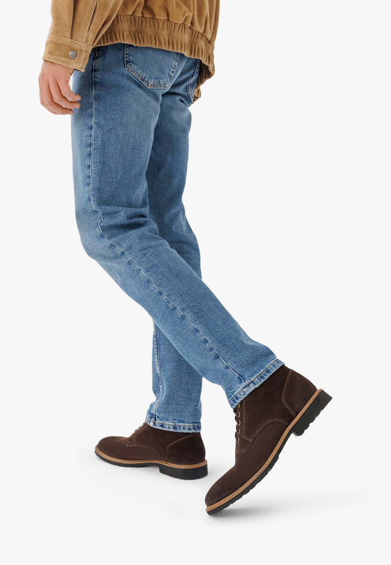 Stivali ankle in suede marrone con lacci, indossati con jeans aderenti azzurri. Gli stivali presentano una suola in gomma e un design semplice.