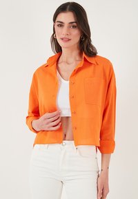 Cropped orange button-up shirt with a front pocket, accompanied by a white tank top, worn over white high-waisted jeans.