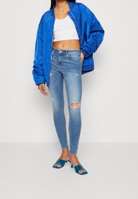 Kvinna som bär en klarblå bomberjacka, vit kort topp, slitna skinny jeans och blå klackskor med öppen tå, står med ena handen i fickan.