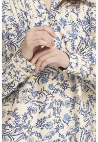 Blouse à motifs floraux en crème avec des fleurs bleues, présentant un col plissé et des poignets boutonnés. Tissu doux avec une finition texturée.