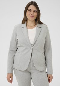 Blazer gris clair avec une coupe ajustée, deux poches latérales et une fermeture à un bouton, fabriqué à partir d'un tissu texturé. Superposé sur un haut blanc.