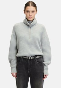 Helles graues Strickpullover mit hohem Kragen und halbem Reißverschluss, kombiniert mit hochgeschnittenen schwarzen Jeans und einem mit Nieten verzierten Gürtel. Weiche Textur und lässige Passform.
