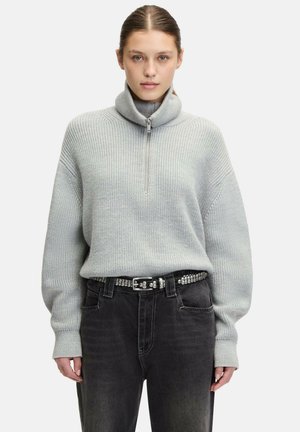 Helles graues Strickpullover mit hohem Kragen und halbem Reißverschluss, kombiniert mit hochgeschnittenen schwarzen Jeans und einem mit Nieten verzierten Gürtel. Weiche Textur und lässige Passform.