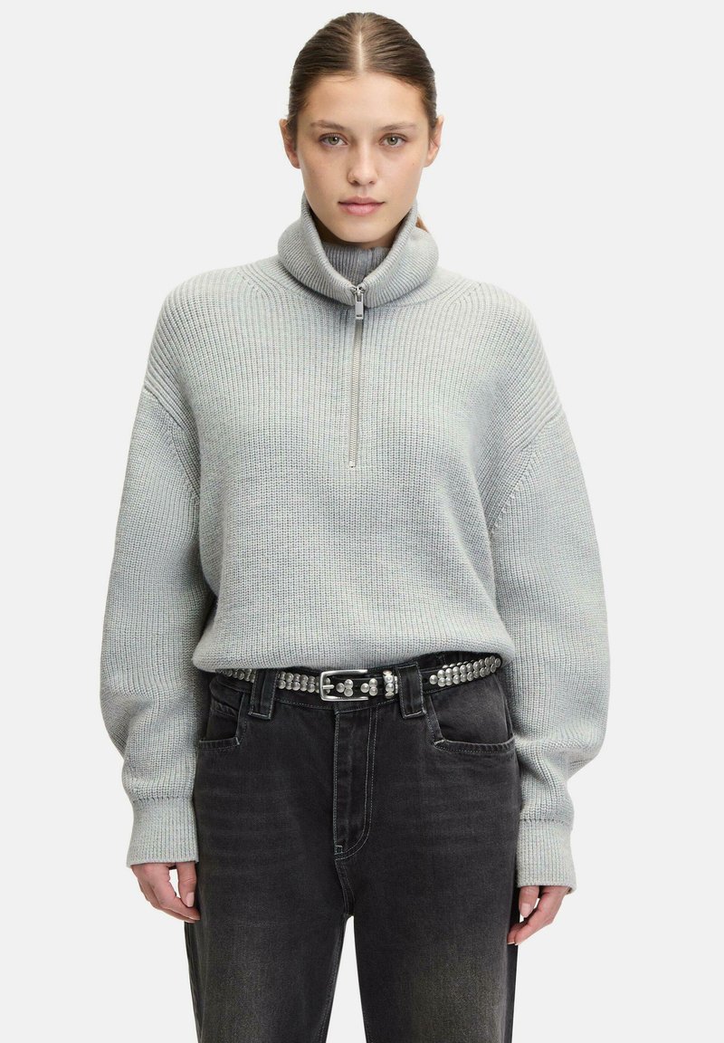 Helles graues Strickpullover mit hohem Kragen und halbem Reißverschluss, kombiniert mit hochgeschnittenen schwarzen Jeans und einem mit Nieten verzierten Gürtel. Weiche Textur und lässige Passform.