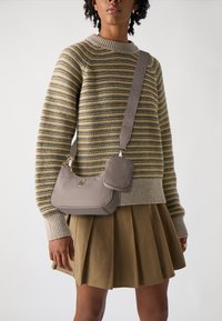 Borsa a mano in pelle beige con forma curvata, dotata di un piccolo pouch rimovibile e di una tracolla testurizzata. Maglione a righe e gonna plissettata.