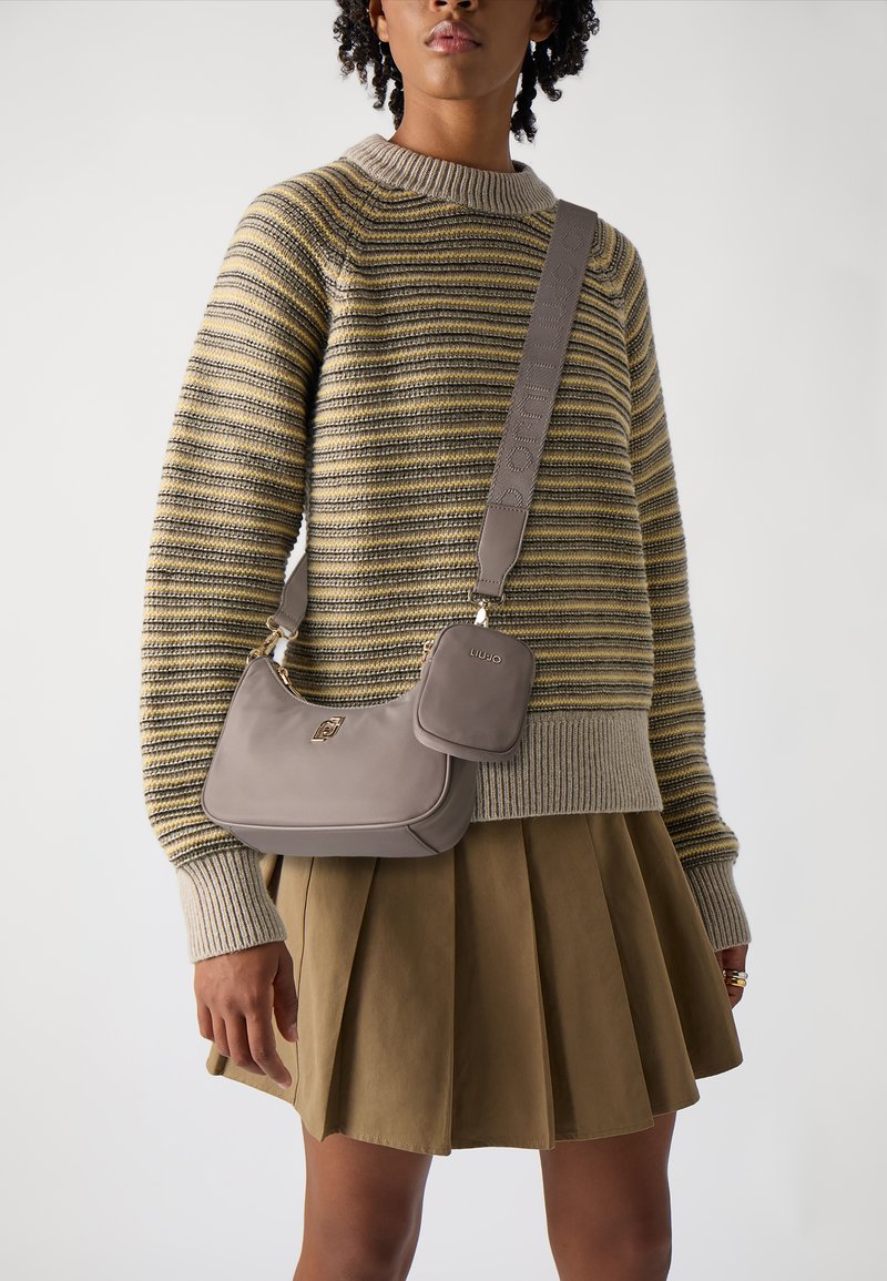 Borsa a mano in pelle beige con forma curvata, dotata di un piccolo pouch rimovibile e di una tracolla testurizzata. Maglione a righe e gonna plissettata.