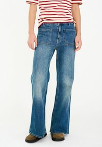 Lichtblauwe spijkerbroek met een uitlopende pijp, voorzien van achterzakken en een lichte vervaging voor textuur. Draagt een rood-wit gestreept shirt.