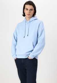 Lys blå hettegenser laget av mykt stoff, med kengurulomme og snor i hetten. Kombinert med mørk denimjeans.