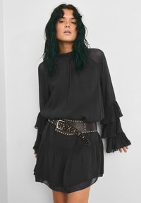 Rochie neagră cu mâneci ondulate, tiv în straturi și un centură la talie cu bolțuri. Materialul transparent și designul cu guler înalt adaugă textură și detaliu.