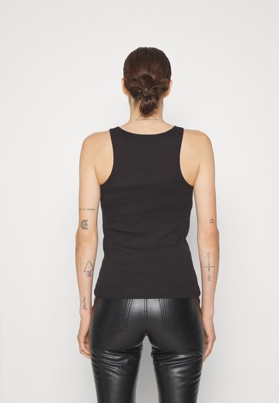 Black ribbed tank top with a round neckline and narrow shoulder straps, paired with glossy black fitted pants. Tattoos visible on arms.
