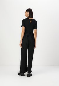 Combinaison noire avec un haut ajusté, des manches courtes et une taille à nouer. Présente une découpe en trou de serrure dans le dos et un pantalon à jambes larges. Texture de tissu lisse.
