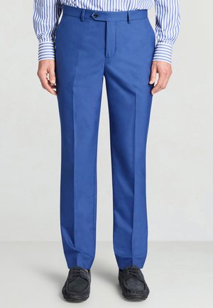 Blaue formelle Hose, getragen mit schwarzen Loafers und einem blau-weiß gestreiften Langarmhemd, stehend vor einem einfarbigen Hintergrund.