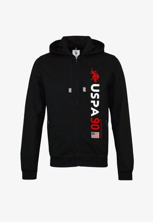 Felpa nera con zip e tasche frontali, con scritta e logo "USPA 90" in bianco e rosso. Indossata con jeans blu e sneakers scure.