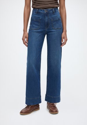 Personne portant un jean bleu taille haute avec de grandes poches avant, des revers de pantalon, une chemise rayée rentrée et des chaussures marron à lacets.