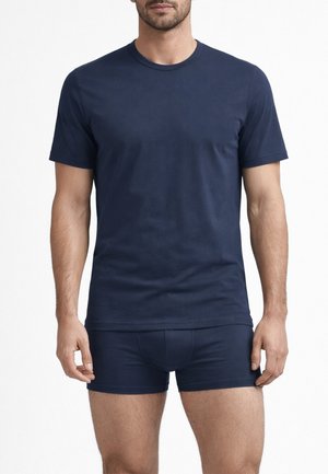 Uomo che indossa una maglietta a maniche corte blu navy semplice e boxer coordinati, in piedi contro uno sfondo bianco.