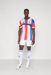 Πολύχρωμη φανέλα ποδοσφαίρου με κόκκινες, μπλε και λευκές ρίγες, διαθέτει στρογγυλή λαιμόκοψη, λογότυπο Adidas και σήμα της Bayern Munich. Φοριέται με λευκά σορτς και κάλτσες.