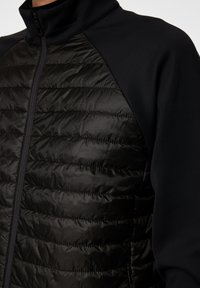 Nära bild av en person som har på sig en svart jacka med en vadderad, quiltad del fram och en slät, svart tyg på ärmarna.