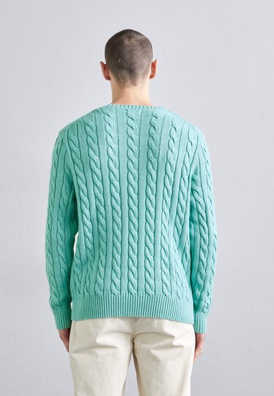 Pull en maille torsadée vert menthe avec un col rond, présentant un motif texturé et des poignets et un ourlet côtelés, porté sur un pantalon crème.