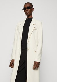 Blazer lung crem cu un design adaptat, rever largi și două butoane. Purtați peste un articol vestimentar negru, completat de ochelari de soare închiși la culoare.